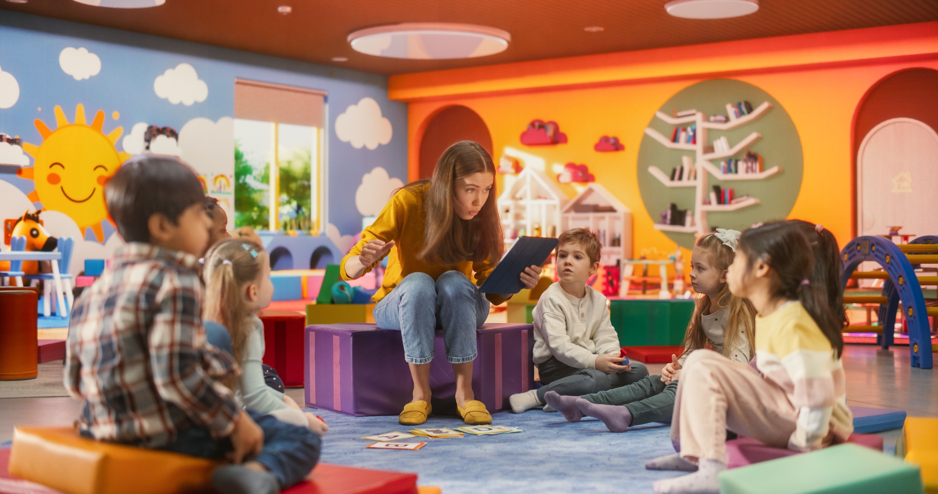 Beautiful Daycare Teacher Using a Tablet Computer to Tell an Educating Story for Young Talented Kids. Female Educator Having Fun Supervising Diverse Group of Small Boy and Girls